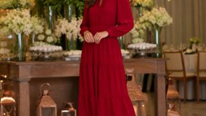 Mulher elegante vestindo vestido longo vermelho em ambiente iluminado com decoração de festa ao fundo.