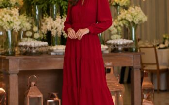 Mulher elegante vestindo vestido longo vermelho em ambiente iluminado com decoração de festa ao fundo.
