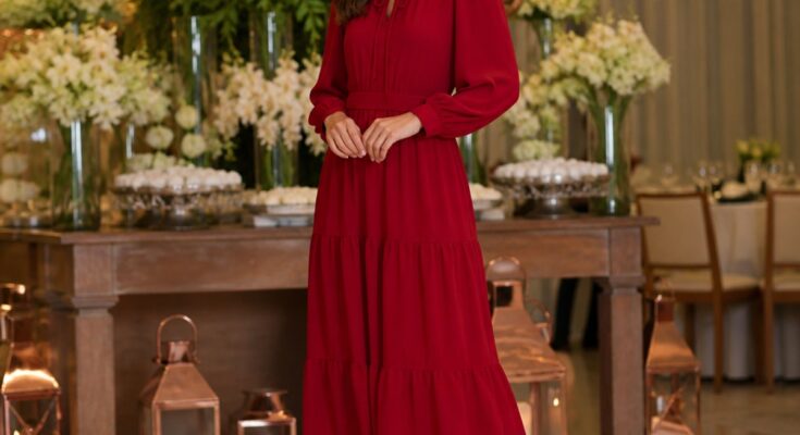 Mulher elegante vestindo vestido longo vermelho em ambiente iluminado com decoração de festa ao fundo.