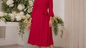 Vestido de festa vermelho em uma mulher elegante, em ambiente iluminado com flores ao fundo, pose confiante.