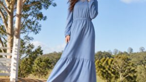 Mulher usando vestido longo azul serenity e bota de couro marrom em ambiente externo com árvores e céu azul claro.