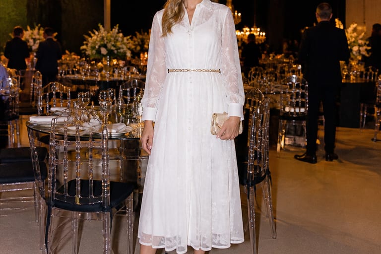 Mulher usando vestido branco transparente com acessórios elegantes em um evento social, em ambiente bem iluminado.