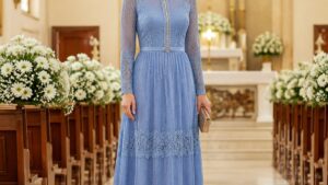 Mulher elegante em pé, usando um vestido longo azul serenity com detalhes em renda e um cinto delicado, em um ambiente de igreja bem iluminado.