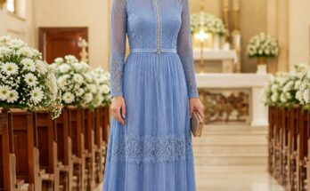 Mulher elegante em pé, usando um vestido longo azul serenity com detalhes em renda e um cinto delicado, em um ambiente de igreja bem iluminado.