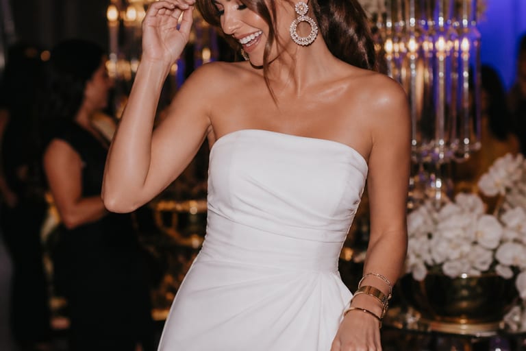 Uma mulher usa um vestido branco em uma festa, cercada por decoração elegante e luzes.