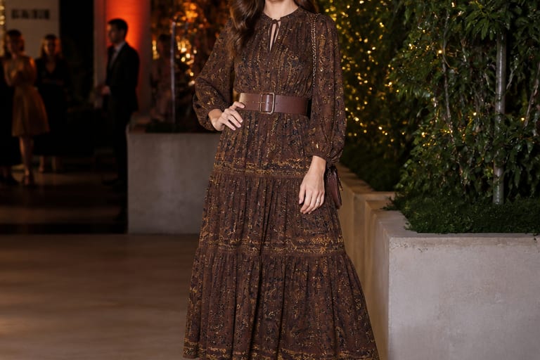 Mulher em vestido longo com cinto e botinhas, posando confiante em evento social.