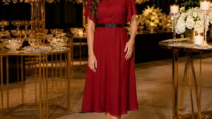 Mulher elegante em vestido de festa vermelho com cinto preto, sorrindo em ambiente iluminado e decorado.