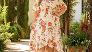 Uma mulher usa um vestido plus size florido em um jardim iluminado, com sapatos de salto alto, transmitindo confiança.