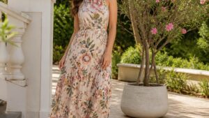 Uma mulher elegante em um vestido longo floral, usando sandálias de tiras, posa descontraída em um jardim iluminado.