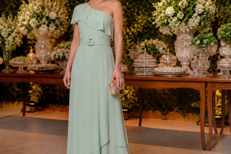 Uma mulher usa um vestido longo com cinto e acessórios, em um ambiente de festa, com sandálias elegantes e iluminação suave.