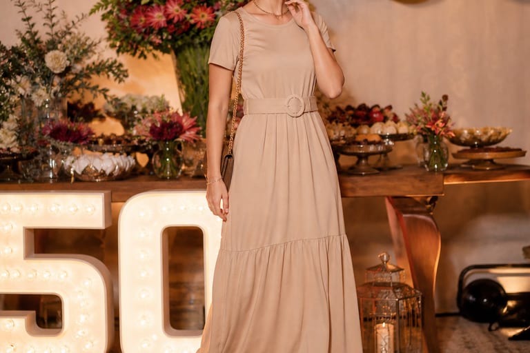Mulher em vestido longo com cinto e acessórios, usando sapatenis, em ambiente festivo.