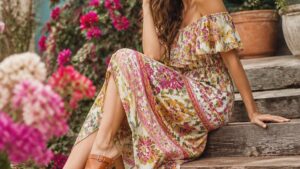 Mulher elegante em vestido floral e tamancos de madeira em ambiente externo com flores ao fundo.