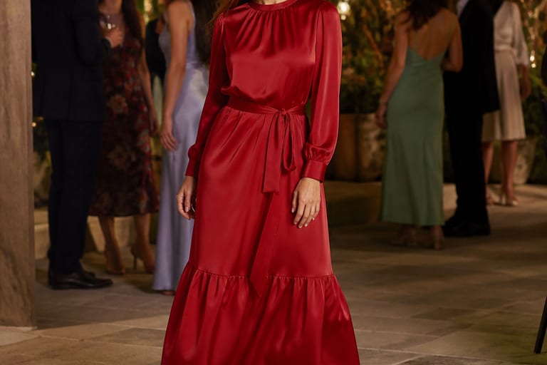 Mulher em vestido longo de seda vermelho e rasteirinhas douradas em ambiente festivo, com convidados ao fundo.